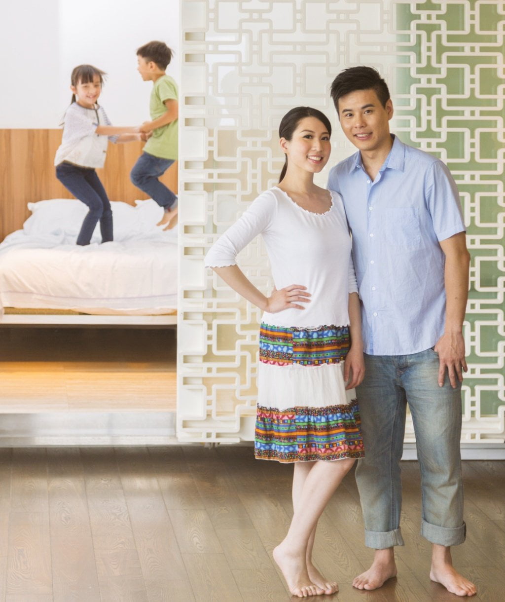 A Chinese family at home. The parents stand together looking at the camera while their two children happily jump on the bed in the background bedroom.