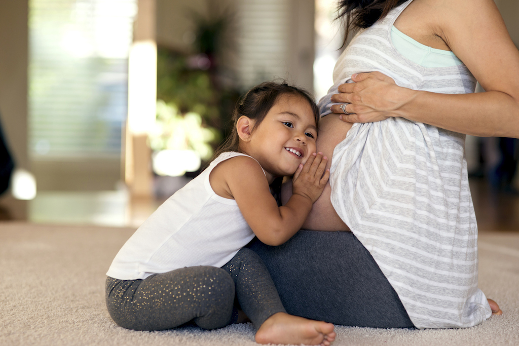 Adorable ethnic child putting her ear to moms pregnant belly